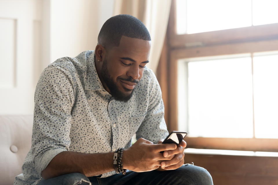 Young african american man holding smartphone texting message or play mobile game sit on sofa at home, smiling millennial black guy using social media app messenger, surfing web on phone indoors