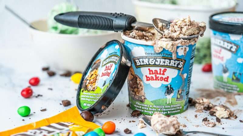 A pint of Ben & Jerry's "half baked" ice cream, with the lid leaning against it and an ice cream scoop laying on top. An open bag of M&Ms is also on the table.