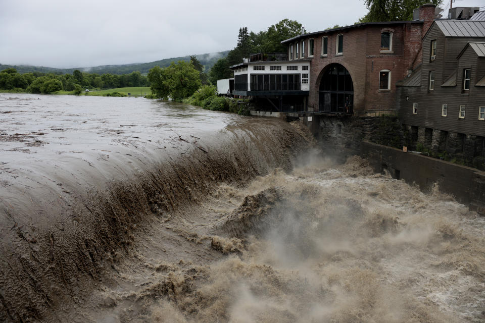 A low dam in a waterway appears to be overflowing with muddy, debris-strewn water next to a few buildings along the bank.