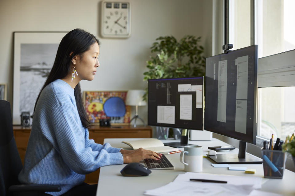 Woman at desk with dual monitors working on a document. She's focused, wearing a casual sweater