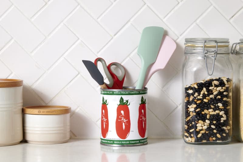 tomato can holding kitchen tool items on a kitchen counter with 2 white containers with wooden lids and a glass of back and white peas next to it. and a tiled backdrop