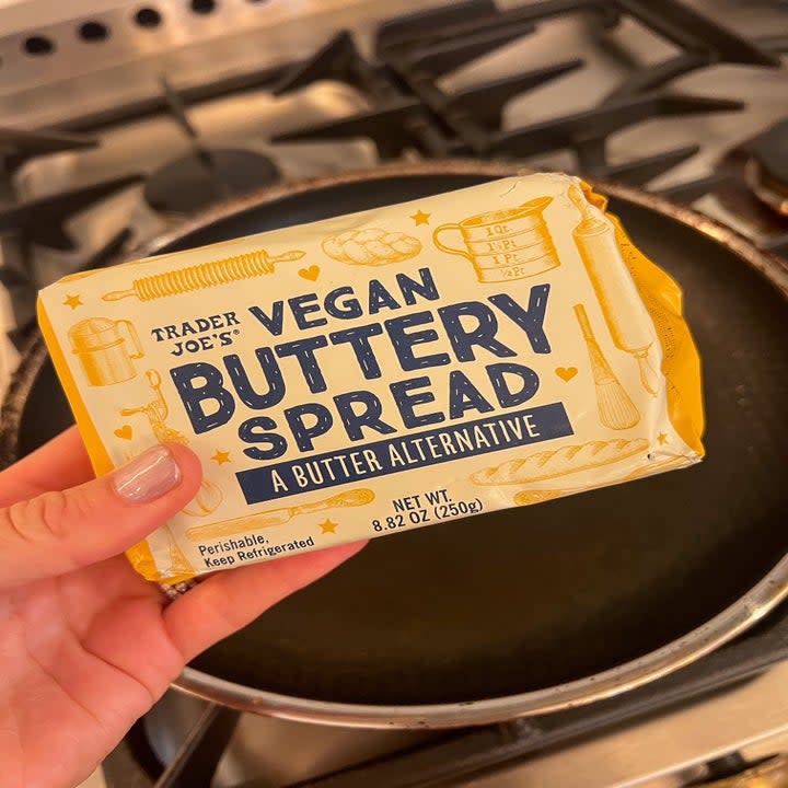 A hand holding vegan butter in front of a skillet.