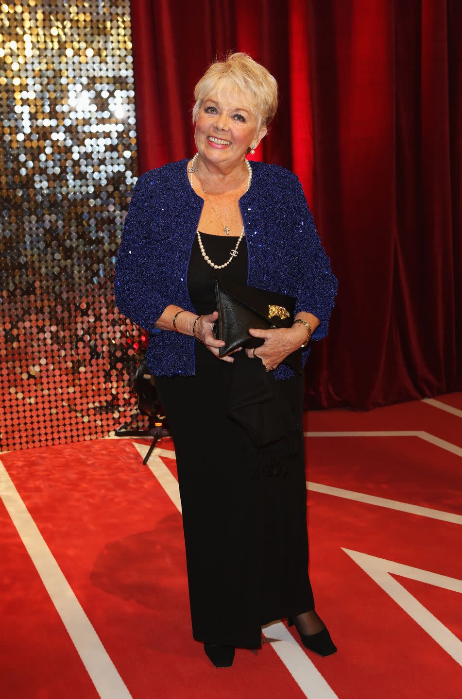 meg johnson, an older lady stands smiling at the camera, she has short light hair, she wears a black dress with blue sequin jacket and black heeled shoes