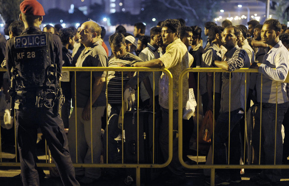 In this photo taken Feb. 9, 2014, a member of an elite police unit, left, watches over migrant workers as they wait for shuttle buses to take them back to their dormitories after spending their day off in Singapore's Little India District. Singapore needs foreign workers, but it doesn’t want them to overstay their welcomes, and firms get fined when they do. That has created a market for “repatriation companies,” which deny allegations from activists and the United States that they use illegal tactics to expel foreign workers. (AP Photo/Joseph Nair)