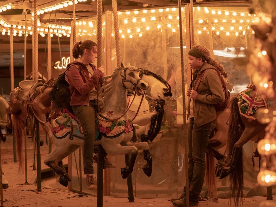 ellie in the last of us sits on a horse on a lit up, dirty carousel. riley is standing in front of her, leaning against another horse, and looking at her skeptically