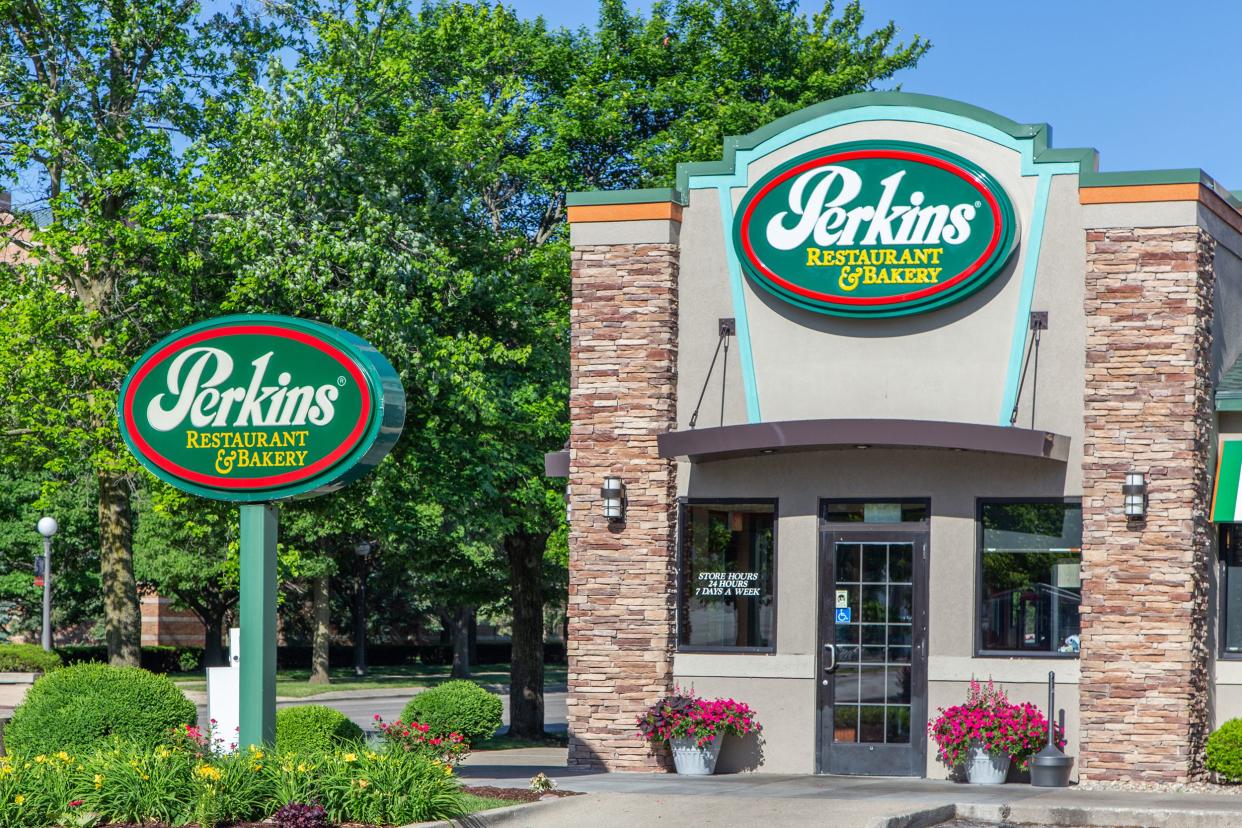 Front exterior of Perkins Restaurant & Bakery in Urbana, Illinois with sign in the foreground, surrounded by shrubbery with trees and blue sky in the background