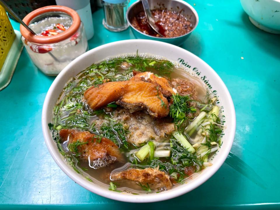 A bowl of fish soup with rice vermicelli noodles and deep-fried fish, dill, spring onions, and served on a blue table