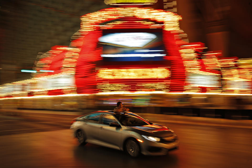 In this April 21, 2020, photo taken with a slow shutter speed, a person takes pictures of temporarily closed casinos from the sunroof of a car in downtown Las Vegas. The usual crowds have been replaced by the occasional bicyclist or driver marveling at the quiet and empty streets. (AP Photo/John Locher)