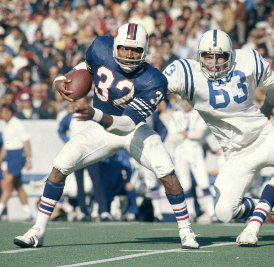 O.J. Simpson on a football field wearing a Buffalo Bills uniform and carrying a football appears to avoid another player.