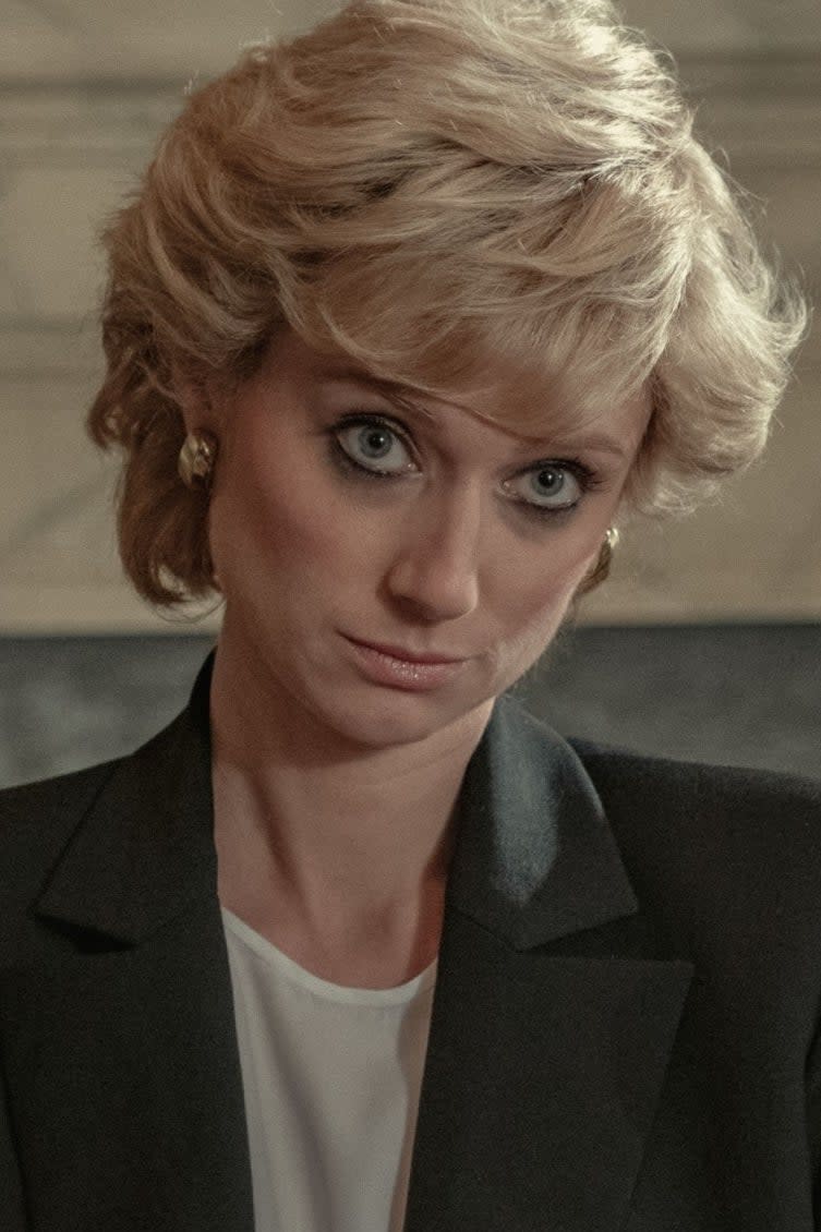 Elizabeth Debicki as Princess Diana, wearing a black blazer over a white top, sits in a formal setting with a serious expression. Scene from The Crown