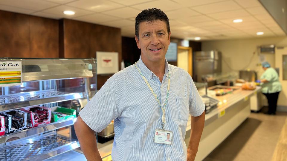 Gavin smiles at the camera with his hands on his hips. He's wearing a short-sleeved shirt with blue stripes on it and has a Jersey government lanyard on. He stands in front of the food counter and there are sachets of condiments. In the background a member of staff prepares some food.