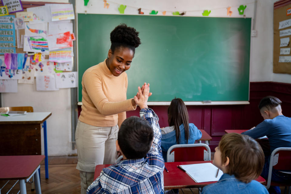 Teacher giving a high-five to a student in a classroom setting. Other students are present