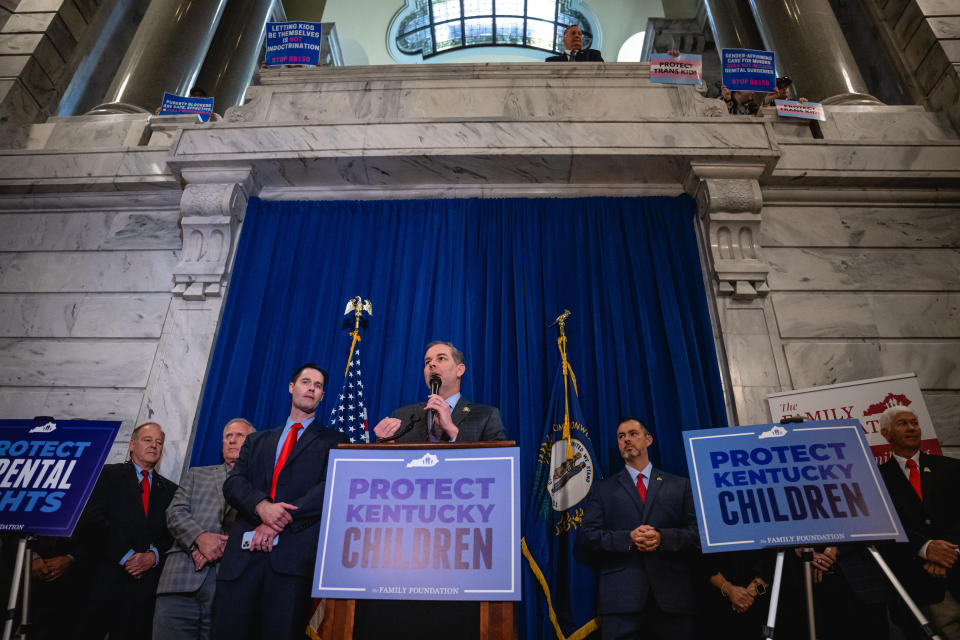 State Sen. Max Wise and other officials at a podium marked Protect Kentucky Children, with protesters above showing such posters as: Protect Trans Kids, Puberty Blockers Are Safe Effective and Fully Reversible and Letting Kids Be Themselves Is Not Indoctrination.