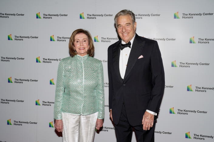 Nancy Pelosi, in white silk trousers and shimmering mint-green evening jacket, and Paul Pelosi, in dinner jacket and bowtie, pose for photos in front of a wall plastered with tape saying: The Kennedy Center Honors.