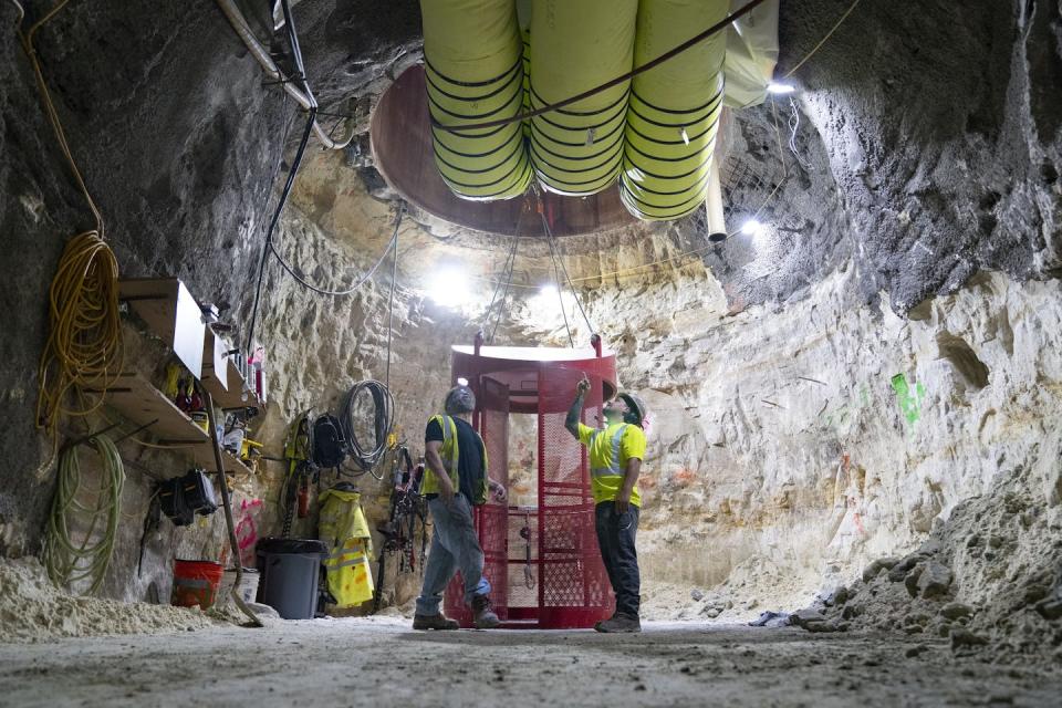 Workers in an underground rock cave look at a giant hole in the ceiling and pipes.