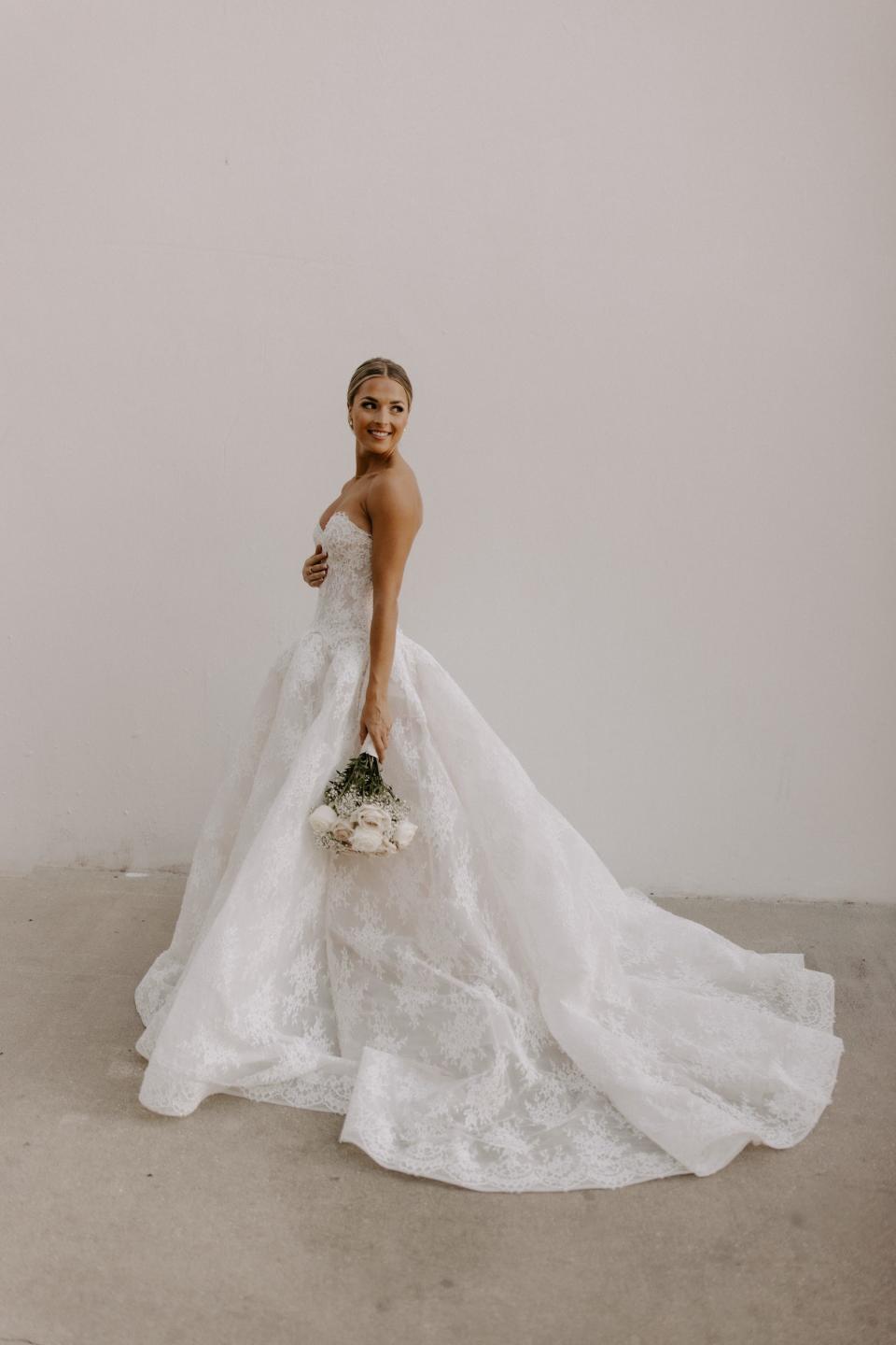 A bride stands in a lace wedding dress with a bouquet of flowers at her side.