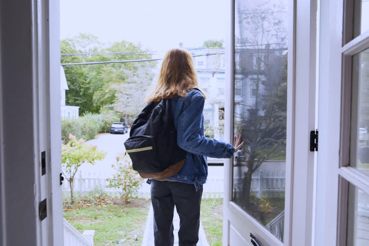 Person with a backpack, wearing a denim jacket, stands in an open doorway looking out at a residential neighborhood