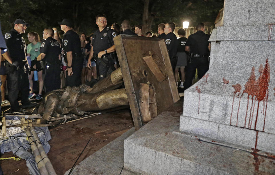 FILE- In this Monday, Aug. 20, 2018 file photo, police stand guard after the confederate statue known as Silent Sam was toppled by protesters on campus at the University of North Carolina in Chapel Hill, N.C. Police are filing charges against three people who they say helped bring down the Confederate statue at North Carolina's flagship university. University of North Carolina Police issued a statement Friday, Aug. 24, 2018, that the department has filed warrants for three people on charges of misdemeanor rioting and defacing a public monument. (AP Photo/Gerry Broome, File)