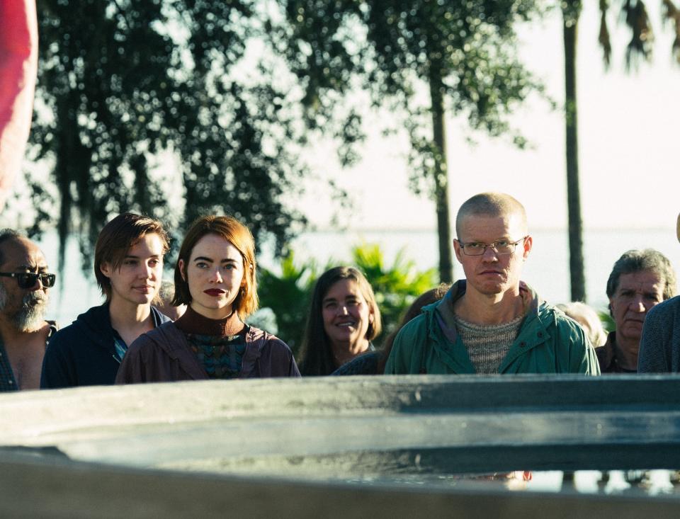 A group of people including Emma Stone and Jesse Plemons stand outdoors behind a concrete structure, looking intently ahead. Trees and sunlight are visible in the background