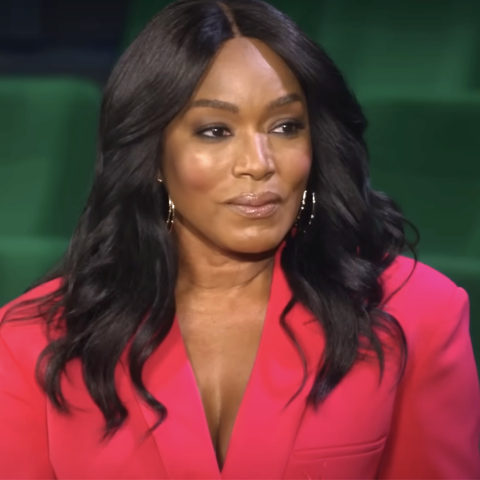 A close-up of Angela in a blazer with hoop earrings, seated against a green background