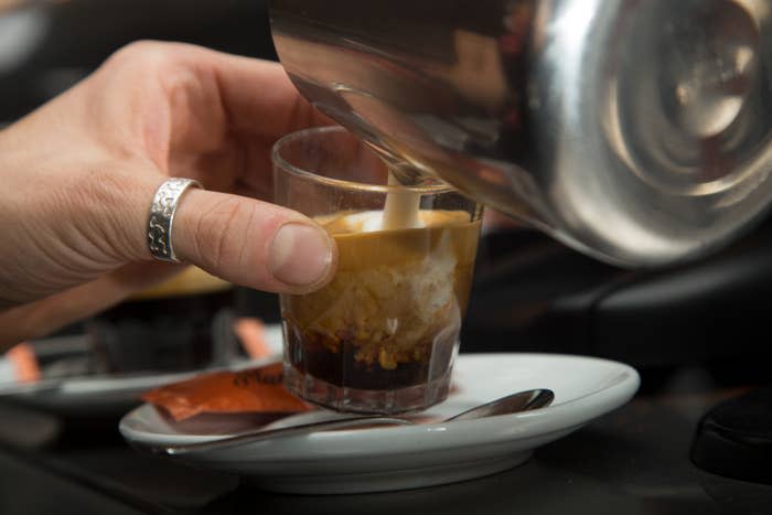 Hand with a silver ring pouring milk into a glass of espresso on a saucer with a spoon and sugar packet