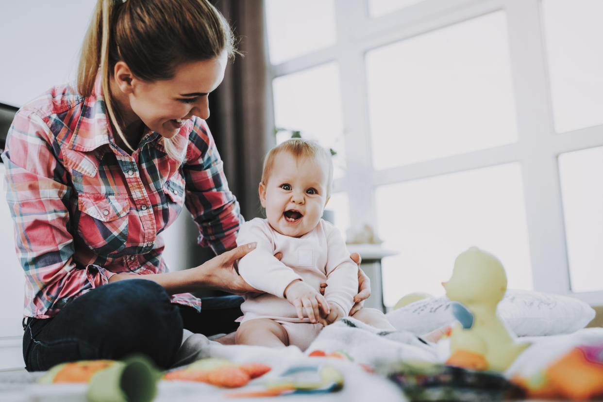 Mother Sits with Child on Floor and Holding Doll. Young Smiling Mother Wears Casual Clothes Playing Toys with Cute Caucasian Baby on Floor near Large Panoramic Window in Living Room.