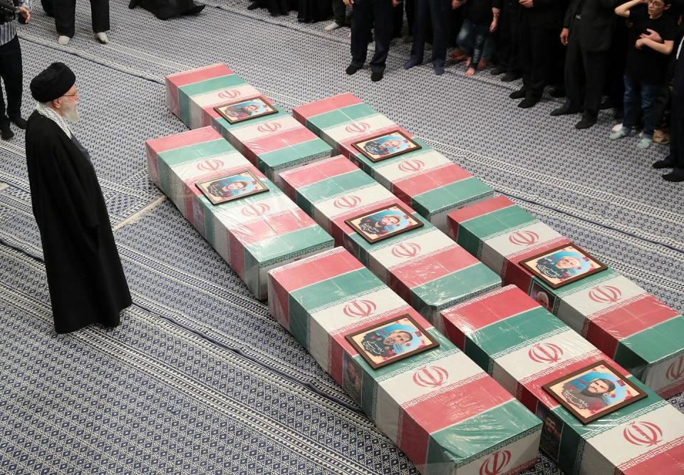 Un hombre vestido con el traje tradicional iraní junto a una fila de ataúdes rojos, blancos y verdes, los colores de la bandera iraní.