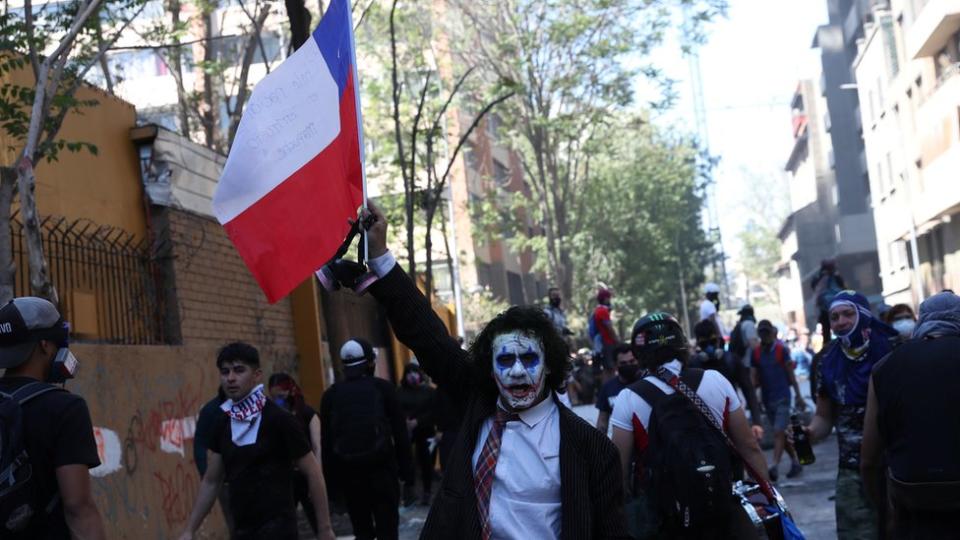 Un hombre con la cara pintada de blanco sosteniendo una bandera de Chile.