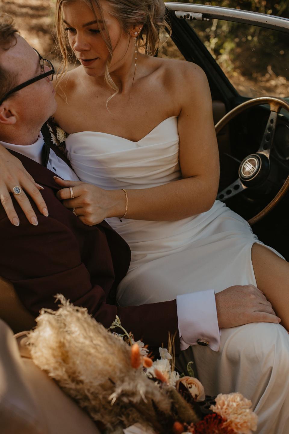 A bride sits on her groom's lap in a car in their wedding attire.