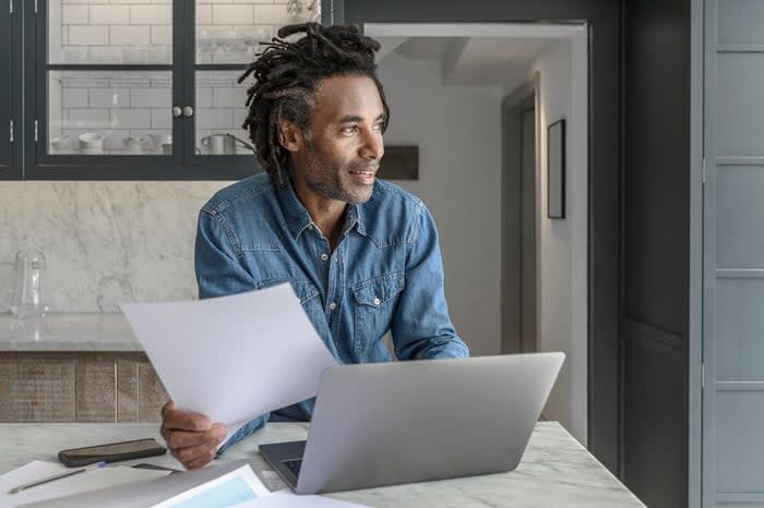 A person looking at paperwork and a laptop while standing at the kitchen counter.