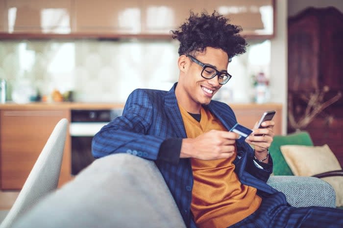 A young man, sitting on a sofa, smiles as he holds a credit card in one hand and looks at his cellphone in another.