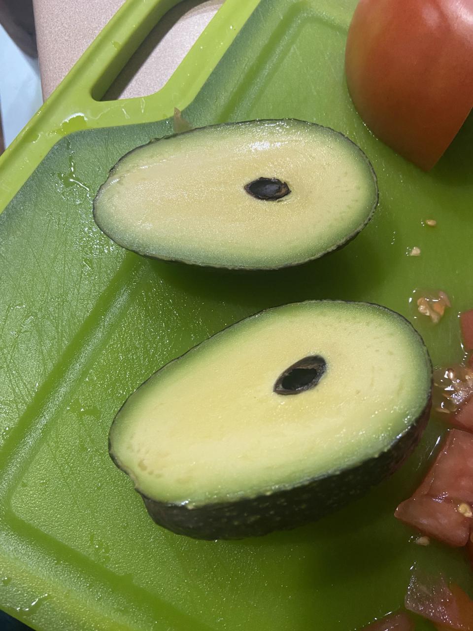 Sliced avocado with a small pit and some chopped tomatoes on a green cutting board