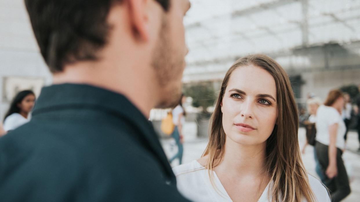  a white woman with long hair is squinting at a man in the foreground of the image, as if trying to remember his face 