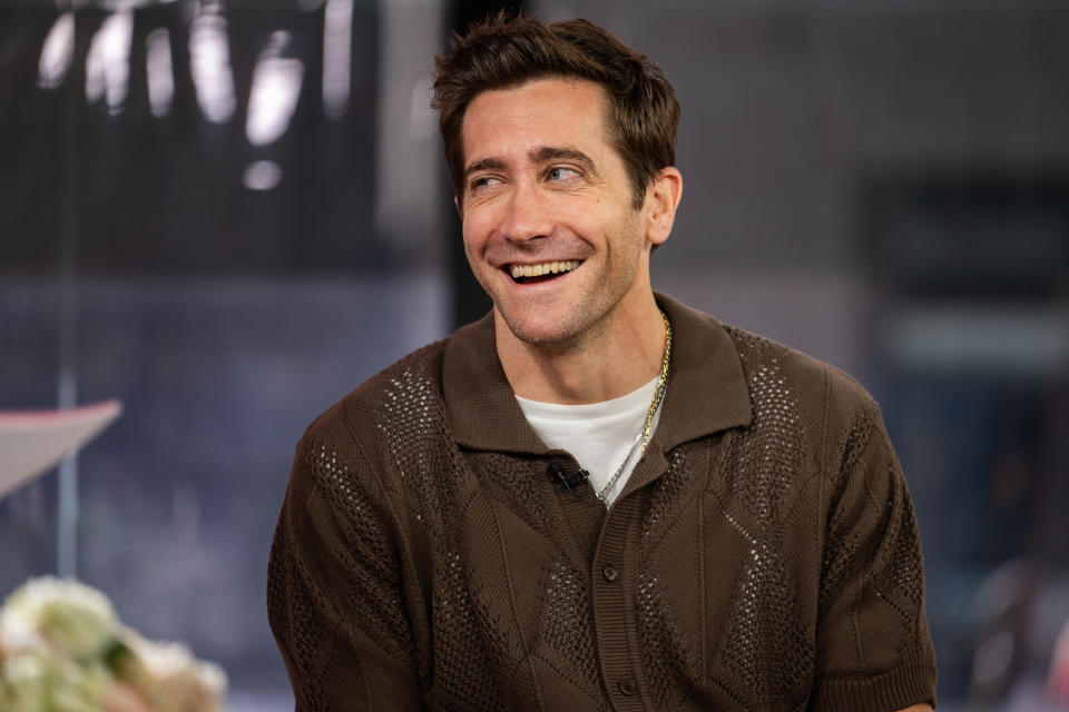 Jake Gyllenhaal is seated and smiling during an interview, wearing a short-sleeved brown knit shirt. The background is a blurred indoor setting