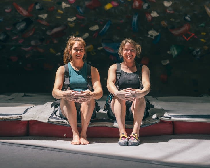 Two women sit on a climbing gym mat smiling at camera. The backdrop is dark but they are bathed in sunlight.