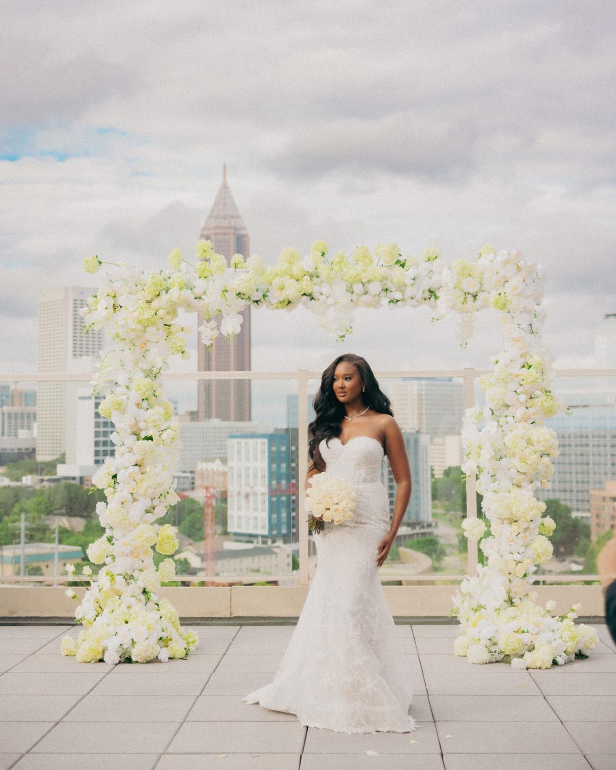 A bride stands in front of a floral archway on a rooftop in her wedding dress.