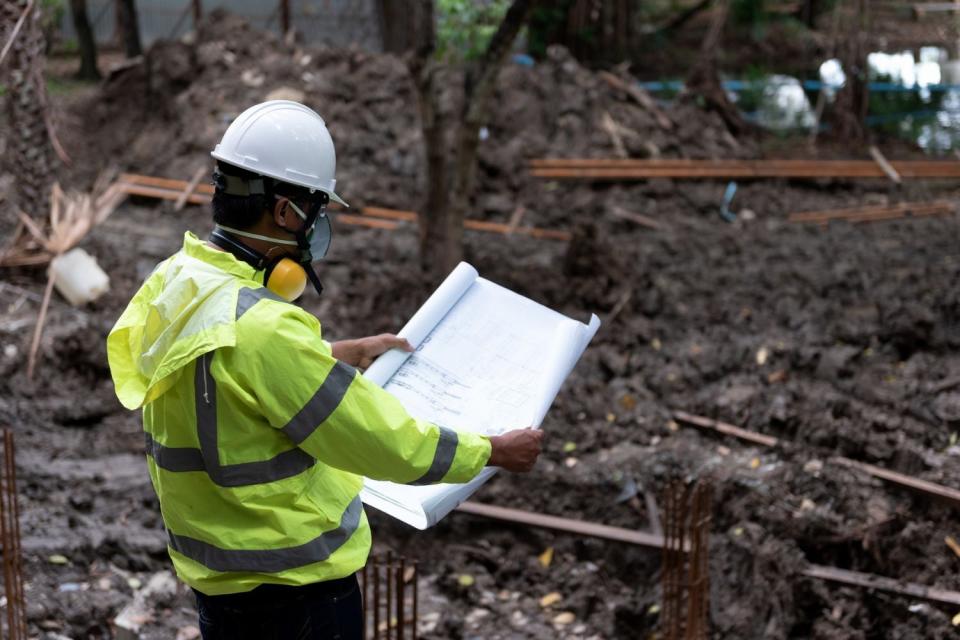 A worker in a bright yellow safety jacket and a white hard hat examines plans on a white sheet of paper while surveying a plot of land.