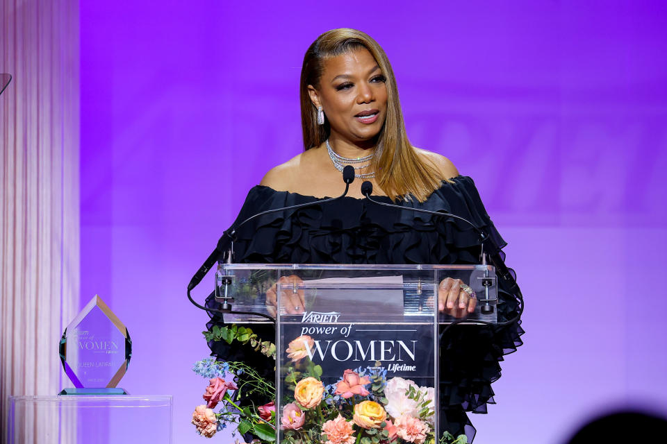Queen Latifah speaking at a podium in an off-the-shoulder dress