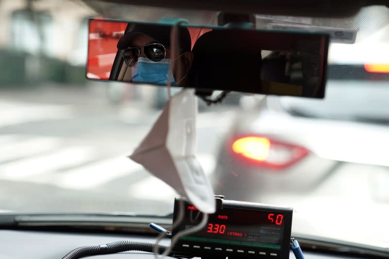 A taxi driver wearing a mask has another mask hanging from his mirror