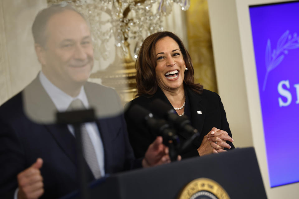 Kamala Harris, wearing a pearl necklace and black suit, smiles and claps at a podium while Doug Emhoff speaks at a formal event