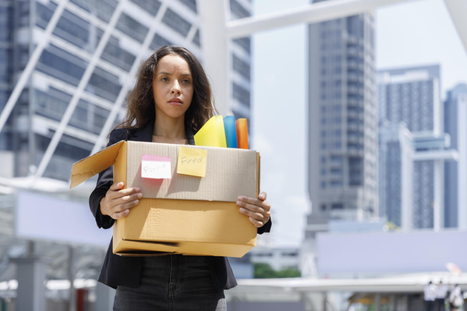 A woman holding a cardboard box with office supplies, including a "Fired" sticky note. She stands in an urban setting with tall buildings in the background