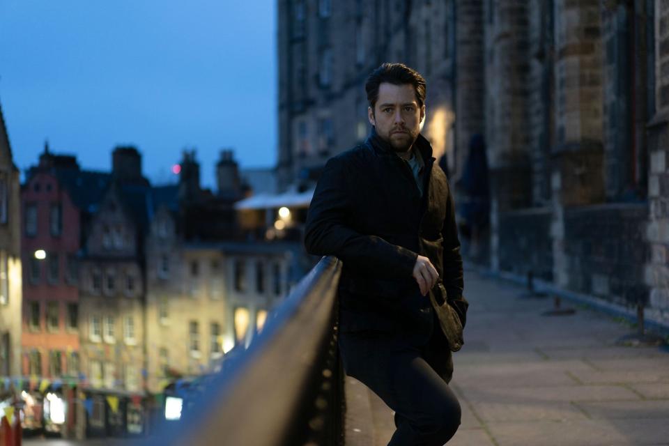 Richard Rankin as John Rebus, wearing a leather jacket and leaning on a wall