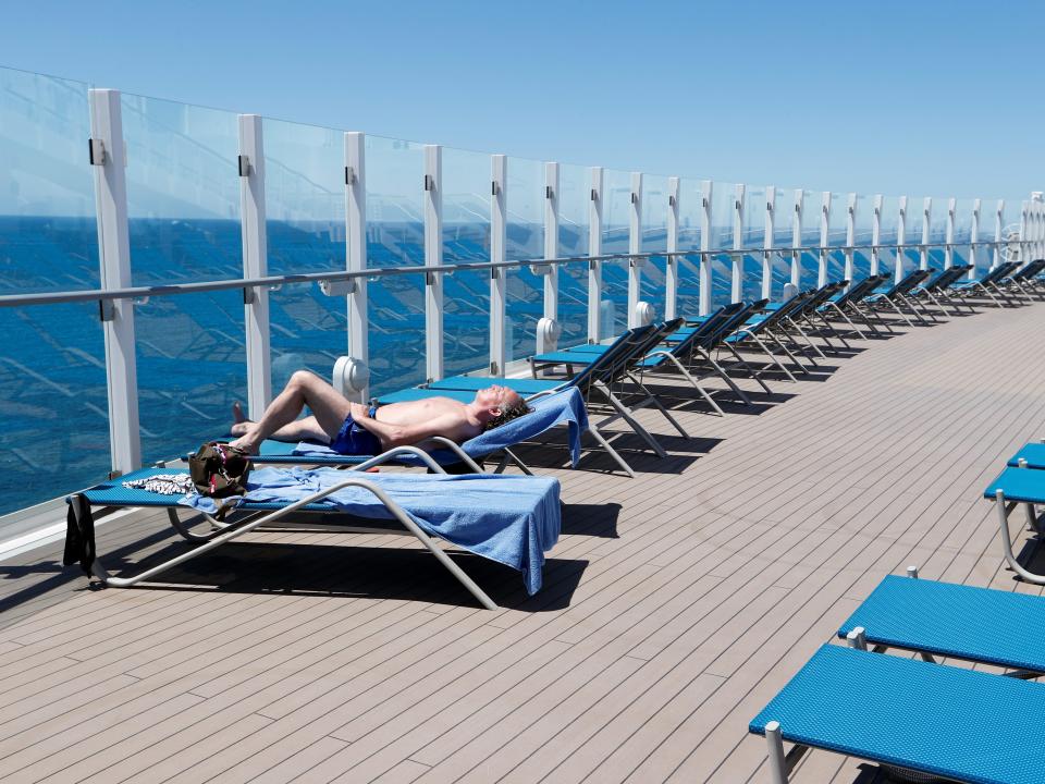 This image shows a line of blue beach chairs on the edge of a cruise ship with a man sunbathing on the left side. It shows the ocean on the far left side and blue skies across the top.