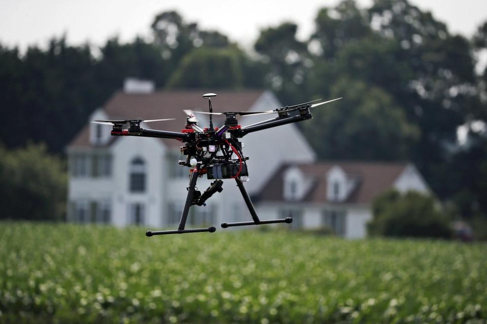A small drone flies over a farmland, with a house in the background.