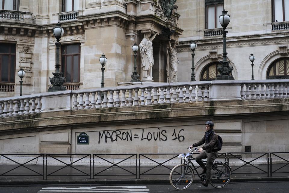 A man rides his bicycle past by a graffiti reading "Macron equal to Louis 16" to reference to the king of France during French Revolution in 1789 on the Paris Garnier Opera house in Paris, Sunday, Dec. 2, 2018. A protest against rising taxes and the high cost of living turned into a riot in the French capital Saturday, as activists caused widespread damage and tagged the Arc de Triomphe with multi-colored graffiti during clashes with police. (AP Photo/Kamil Zihnioglu)