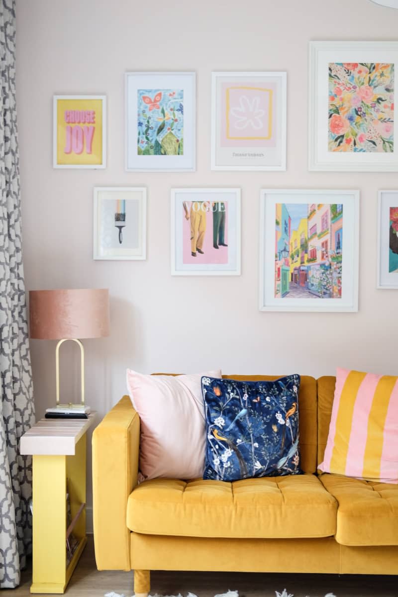 gallery wall above yellow couch with pink and yellow striped and blue throw pillow, slender side table with gold lamp stand and pink lamp shade in corner