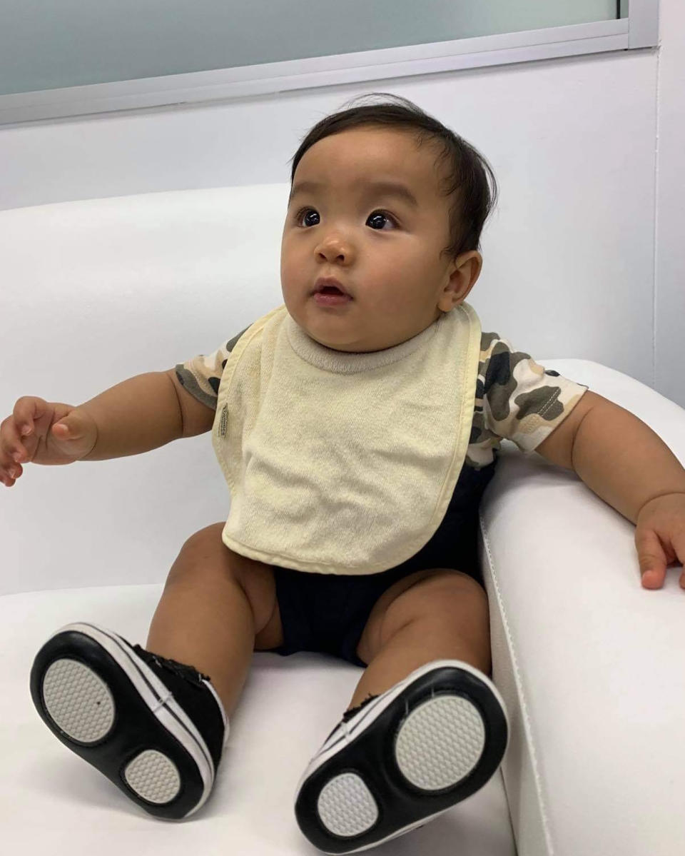 An Asian baby wearing a light yellow big and black shoes looking to the left of the camera.