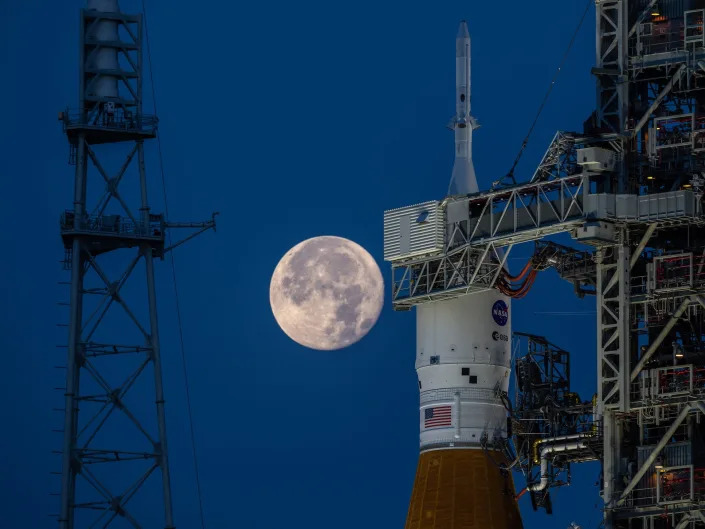 bright moon rises in dark blue sky behind the top of an orange nasa rocket