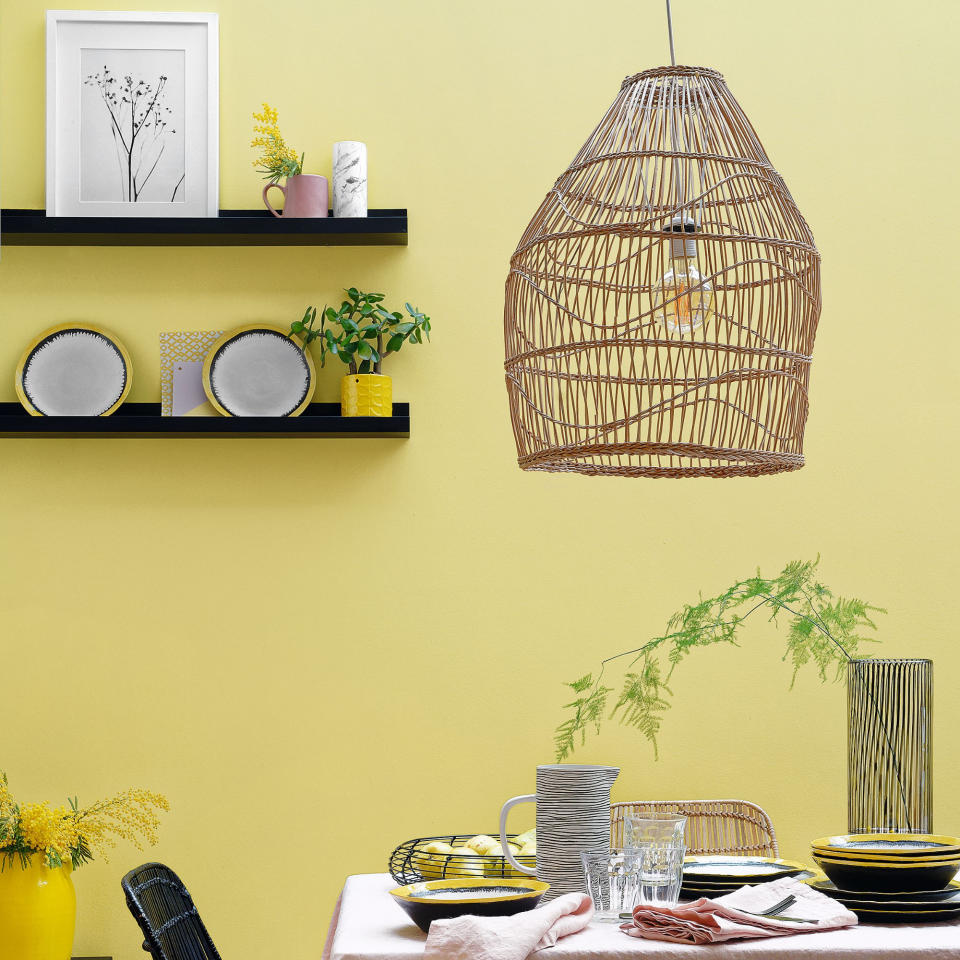 Dining table with pink tablecloth, yellow walls, black shelves and chairs and large wicker lampshade