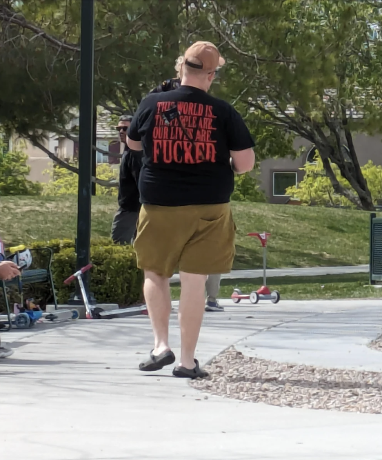 Person in t-shirt and shorts walking at a park, with obscured text on shirt, near children's scooters